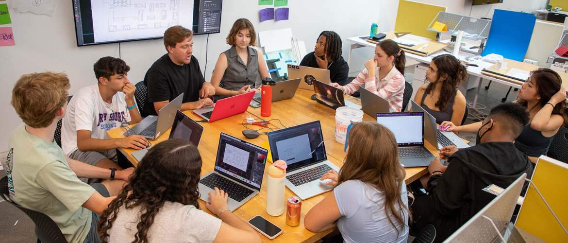 A group of people sitting around a large wooden table with laptops and notebooks, engaged in a collaborative discussion or work session. A large screen on the wall displays a blueprint or architectural drawing.