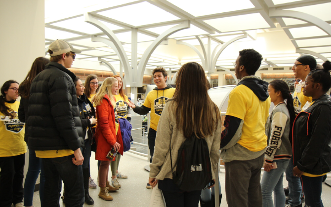 A diverse group of people in yellow t-shirts and other casual clothing stand in a modern building with arched ceilings, some talking and gesturing.