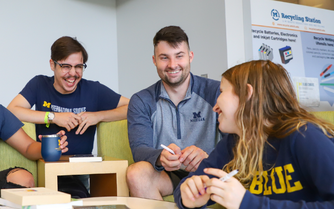Three people in navy blue shirts are sitting on a couch and smiling. One person is holding a blue tumbler. Behind them is a recycling station sign.