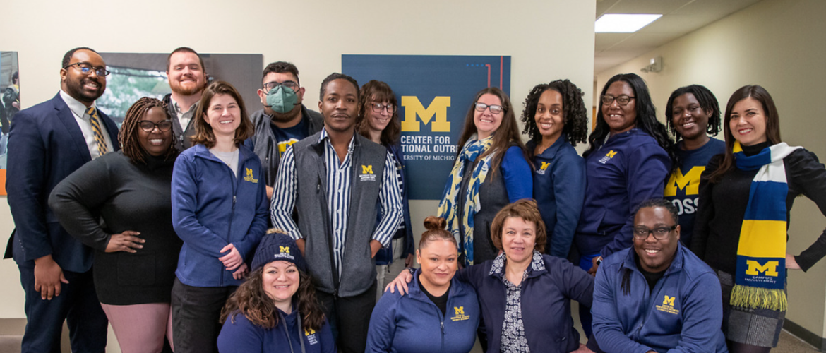 A diverse group of people, some wearing Michigan Wolverines apparel, pose for a photo in front of a 