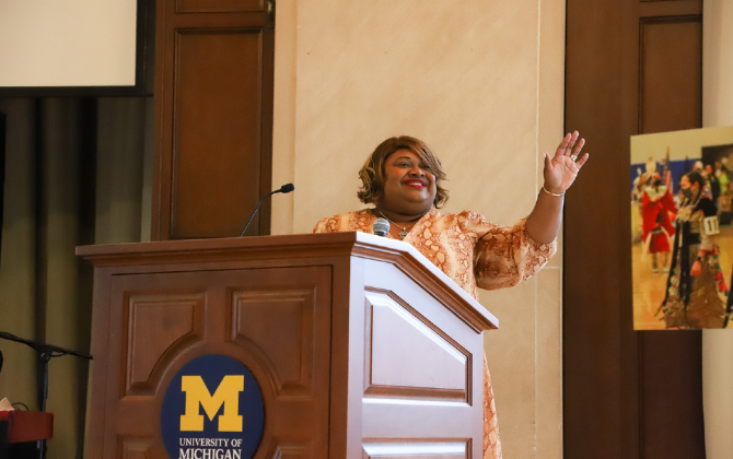 A person stands behind a wooden podium with the University of Michigan logo and waves to an audience.