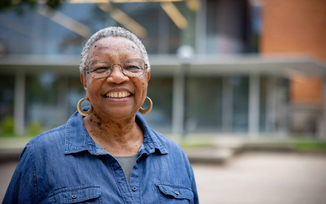 A smiling person wearing glasses and a denim shirt stands in front of a blurred building.