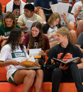 Two students, one in a white and green shirt and the other in a black shirt, sit on bleachers and look at each other while holding papers and books. Other students are visible in the background, also seated on bleachers and looking at papers.