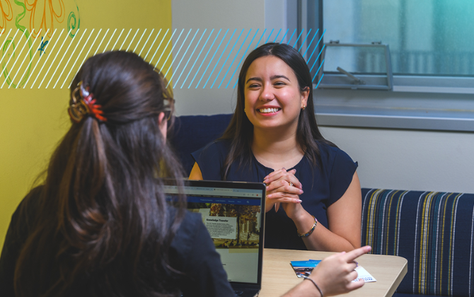 Two individuals are sitting at a table in front of a laptop, with one smiling and gesturing with their hands while the other looks at the screen.