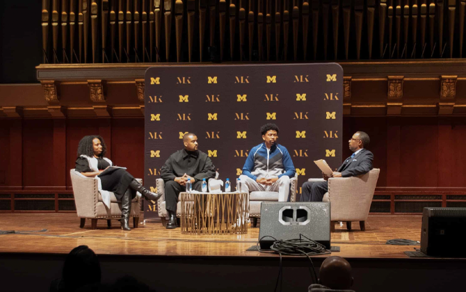 Four people are seated on a stage in front of a backdrop with the letter M repeated. On the left, a woman sits in a chair, holding a notebook. Next to her, a man in a black shirt is seated. In the center, a young person in a blue jacket sits. 