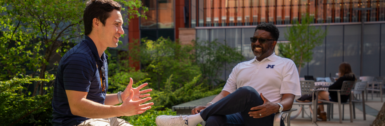 Two individuals are conversing outdoors on a patio area with lush green foliage and modern building architecture in the background. One person wears a navy blue collared shirt and is gesturing with their hands while speaking.