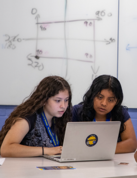 Two individuals are focused on a laptop screen, with a whiteboard displaying mathematical equations and diagrams in the background.