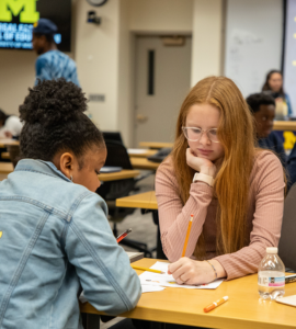 Two students sit at desks in a classroom, one with long red hair wearing glasses and the other with dark curly hair wearing a denim jacket with cartoon characters on the back.