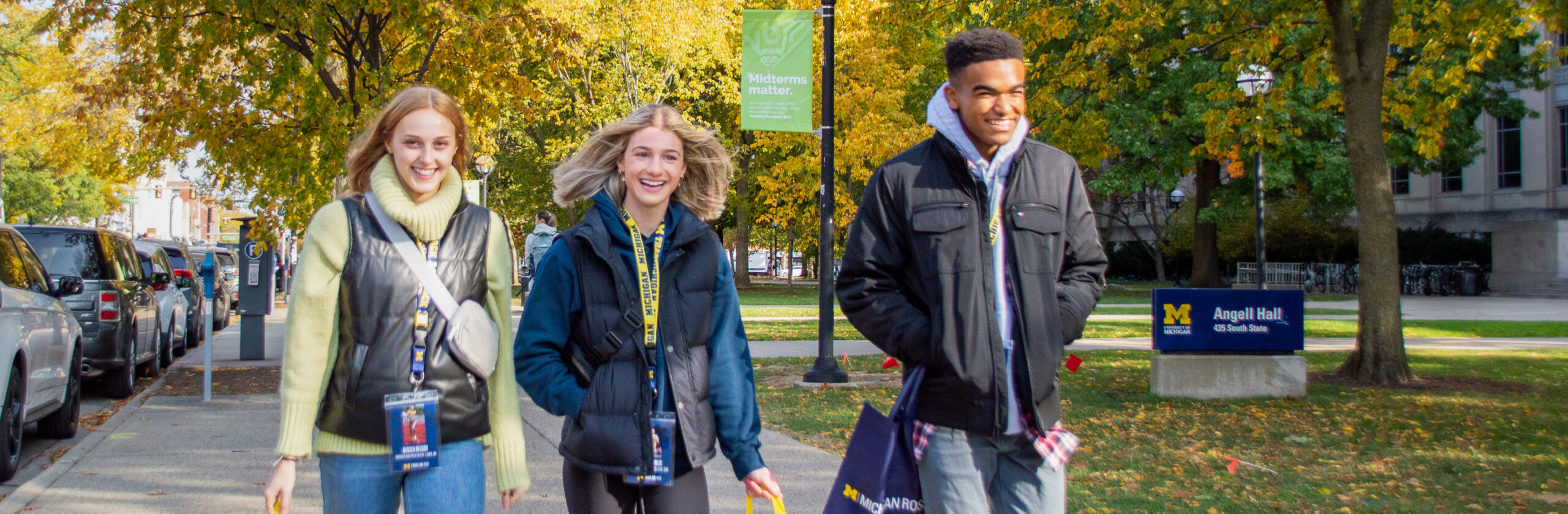 Three people walk along a sidewalk on a tree-lined campus in autumn. The person on the left carries a yellow bag, the middle person carries a yellow bag, and the person on the right carries a blue bag.