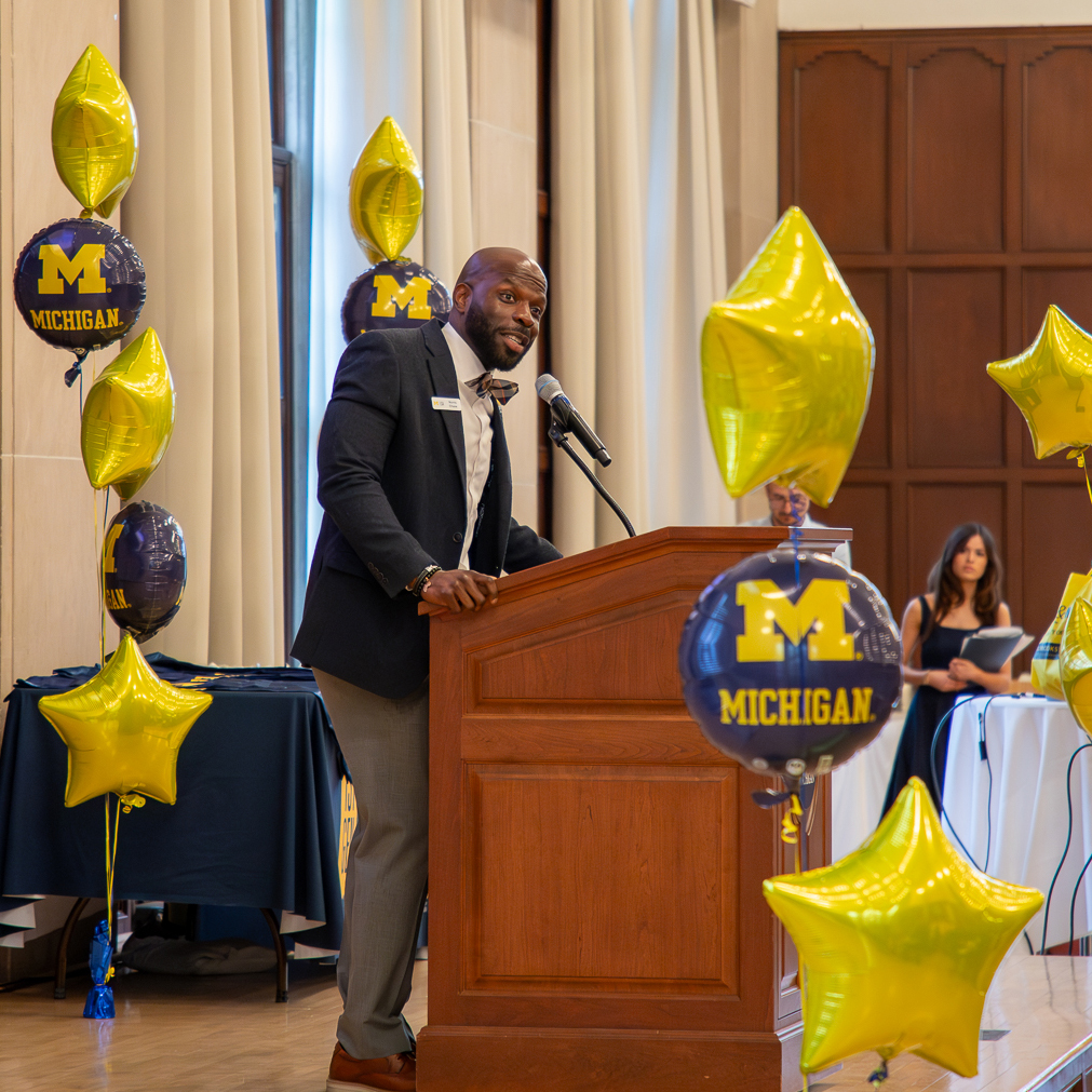 Black male wearing a jacket and tie standing at a lecturn with Michigan balloons surrounding