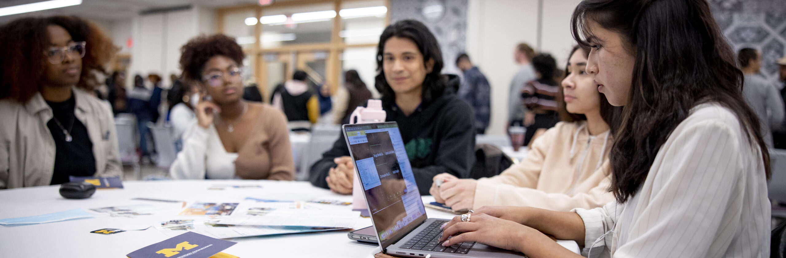 Group of students seated at a table. Girl on far right working on computer.